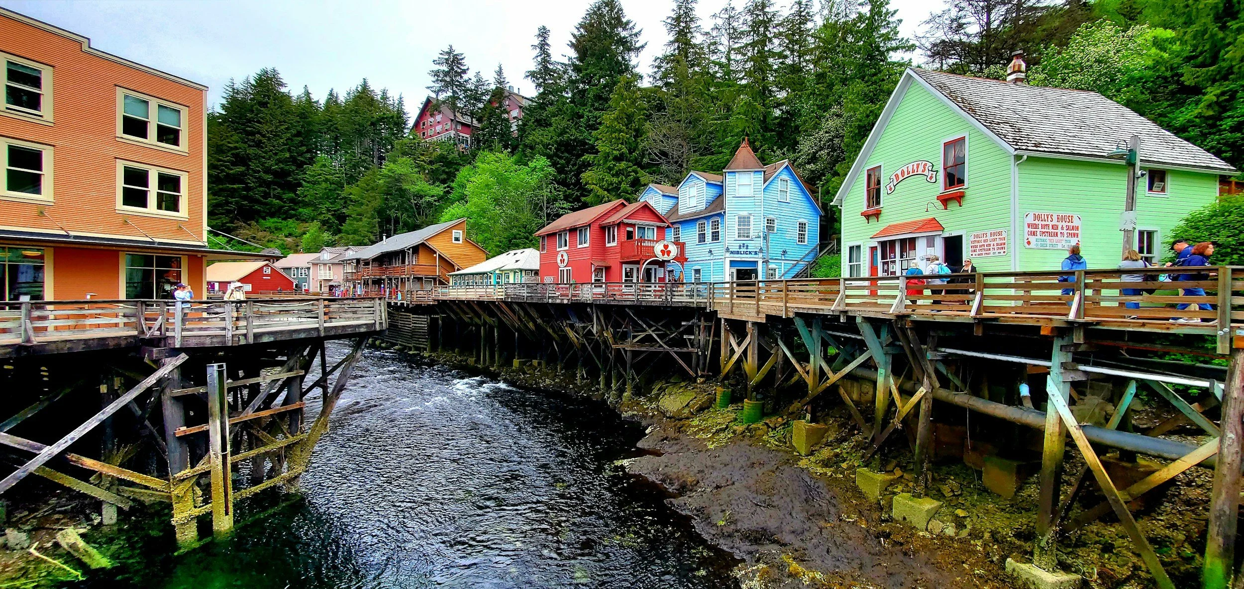 Colorful houses on stilts along a riverwalk with people walking, surrounded by green trees and hillside.