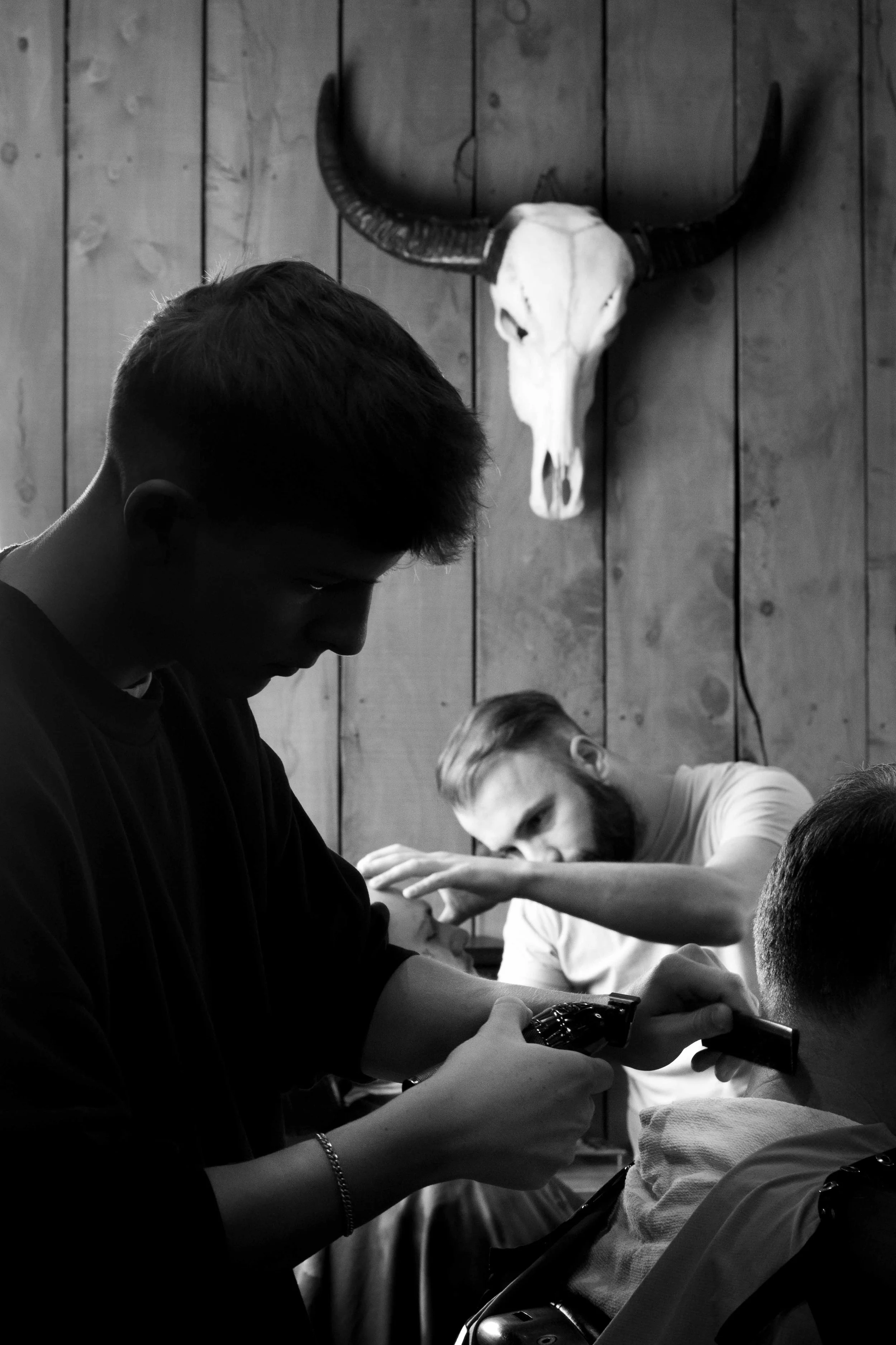 A black and white photo of a barber shaving a man's beard with a straight razor, with a man in the background getting a haircut, while a skull with large horns hangs on a wooden wall.