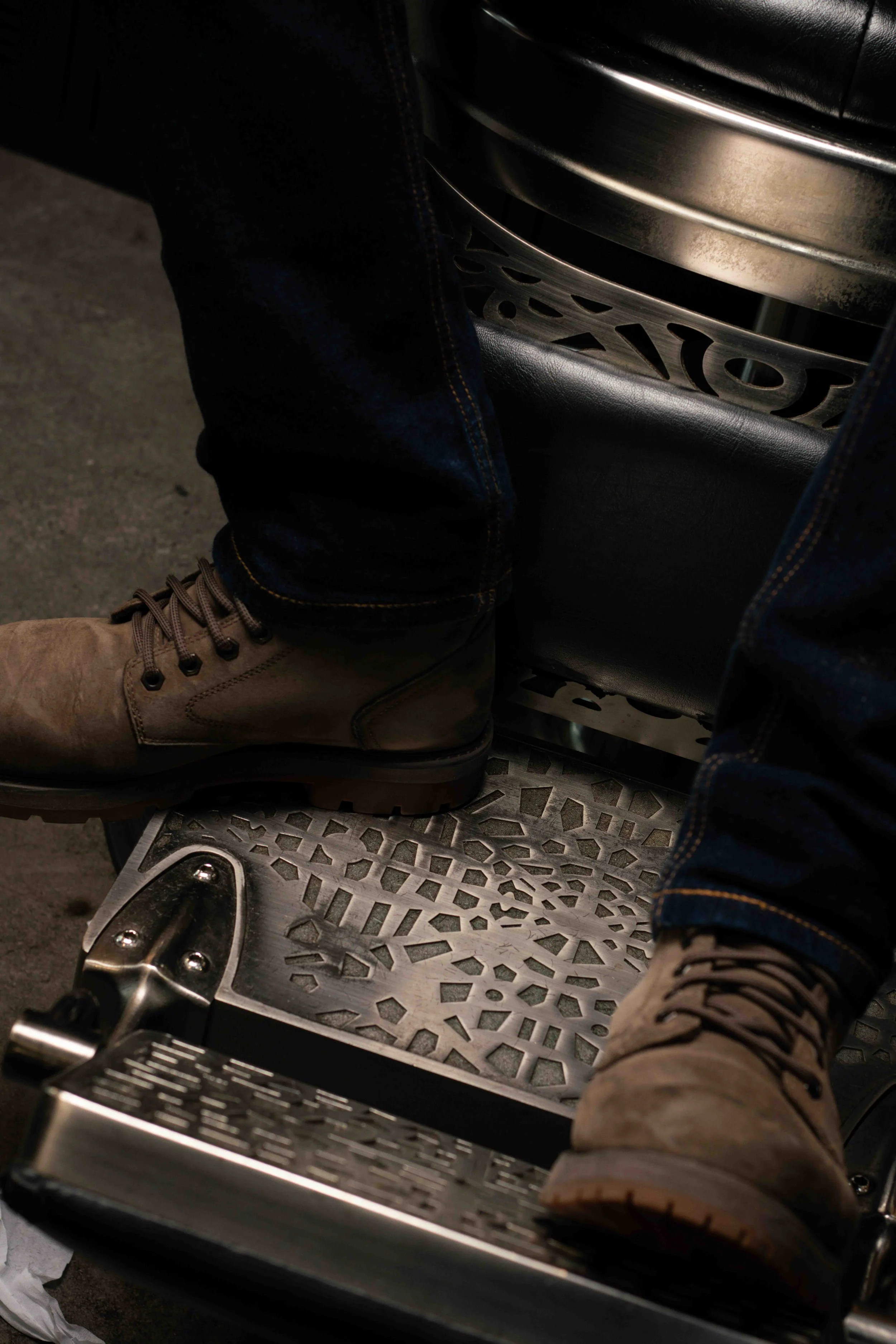 Person wearing tan work boots and blue jeans, standing on a metal platform with a textured surface, next to a piece of equipment with a perforated metal shield and a large metallic object mounted on a black seat.