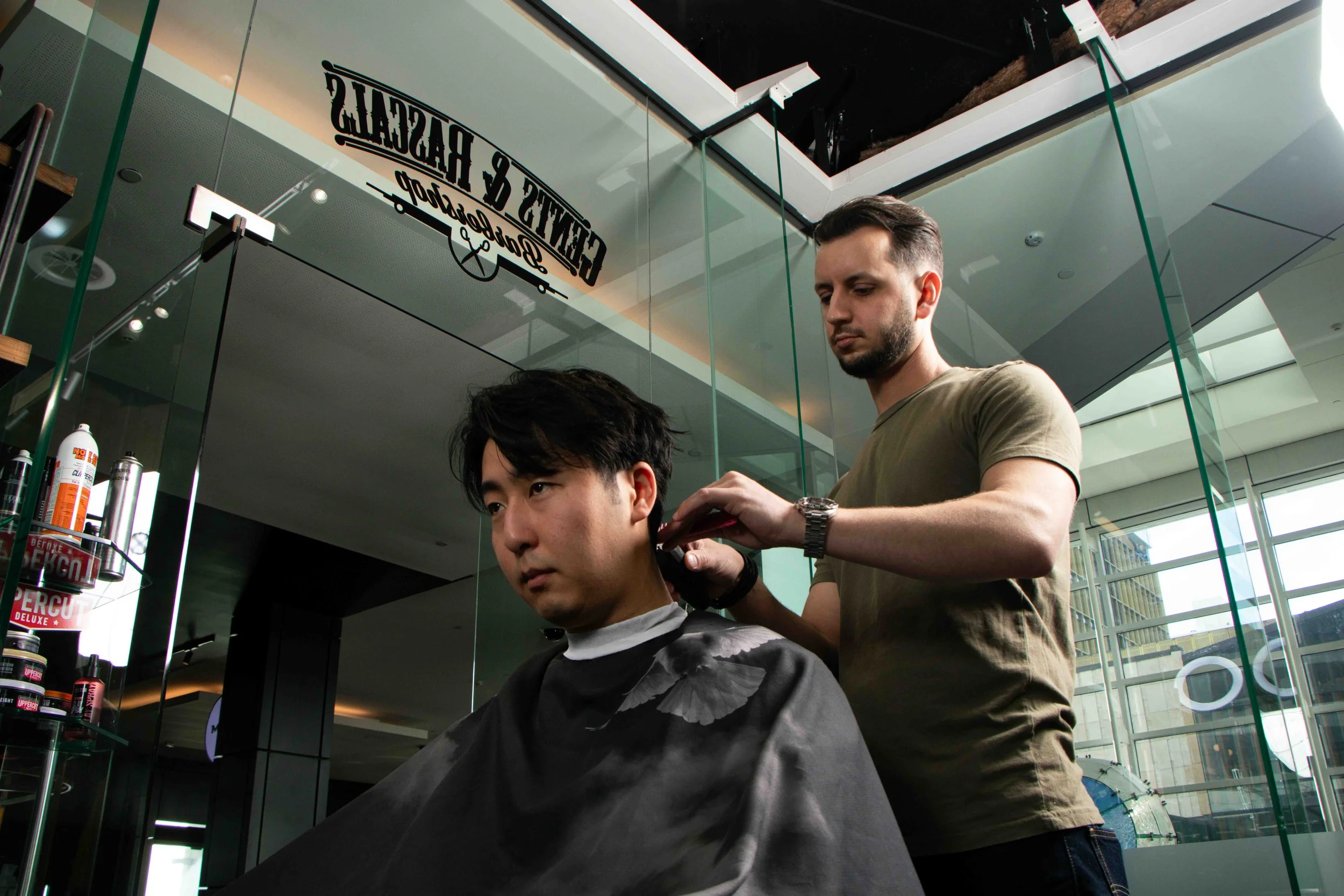 A man getting a haircut at a modern barbershop with glass walls and urban views outside.