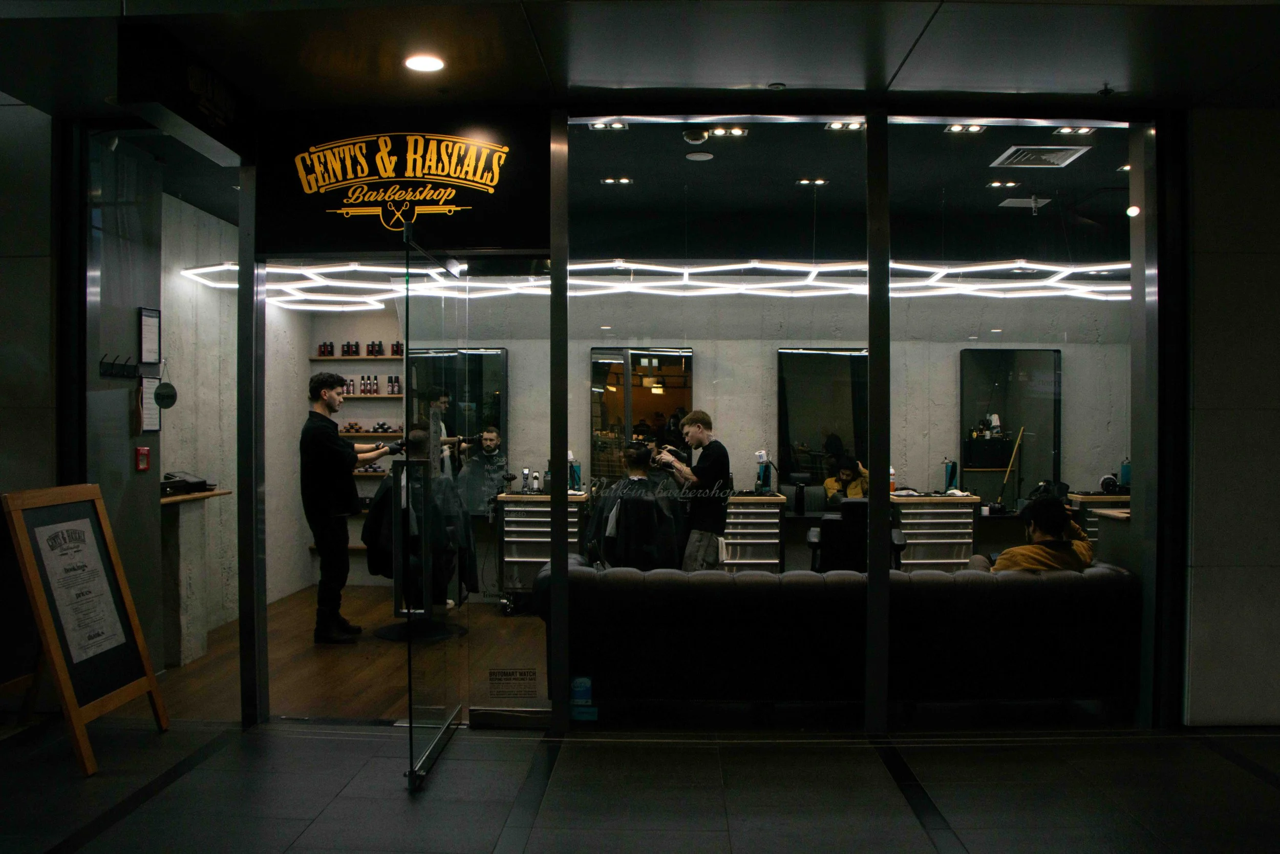 Inside a modern barbershop named 'Gents & Rascals,' with customers getting haircuts and barbers styling hair, viewed through glass windows at night.