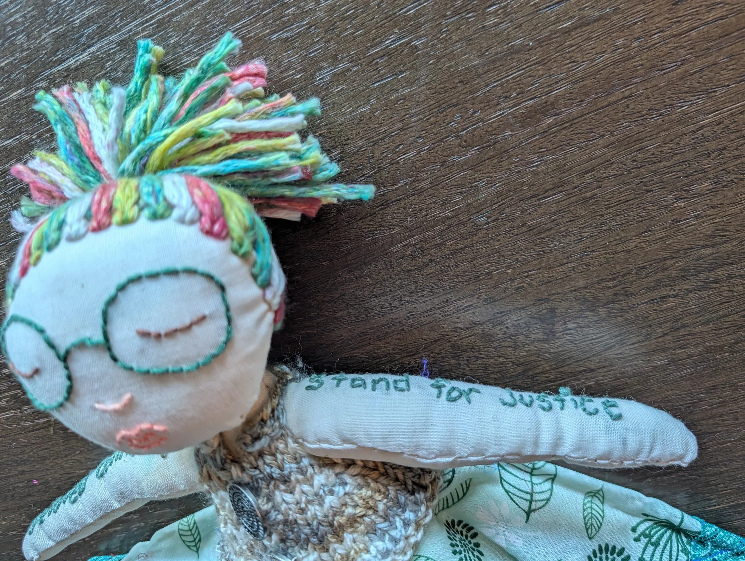 Close up of doll with glasses and red, green,  chartreuse, and white hair in a ponytail. Her visible arm reads "stand for justice"