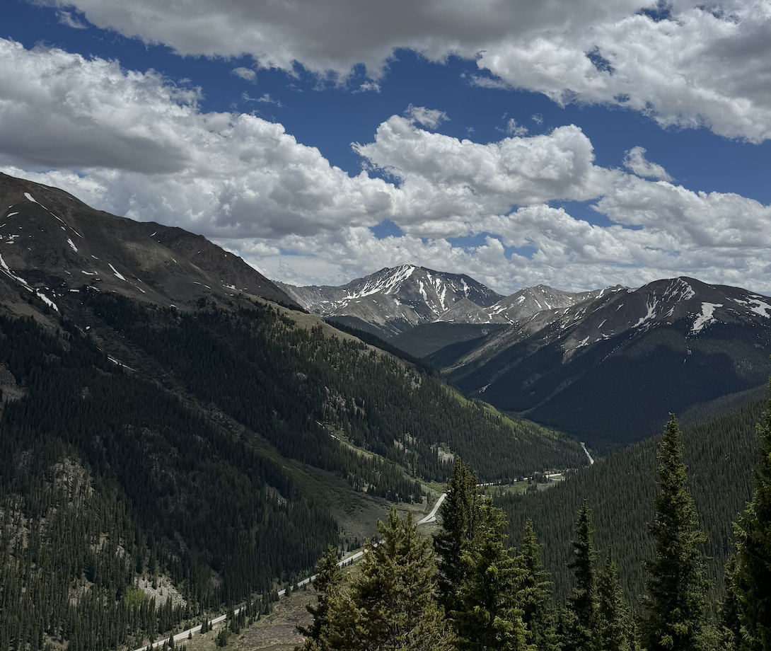 A mountain landscape with forested slopes and snow patches, a winding road, and a partly cloudy sky.