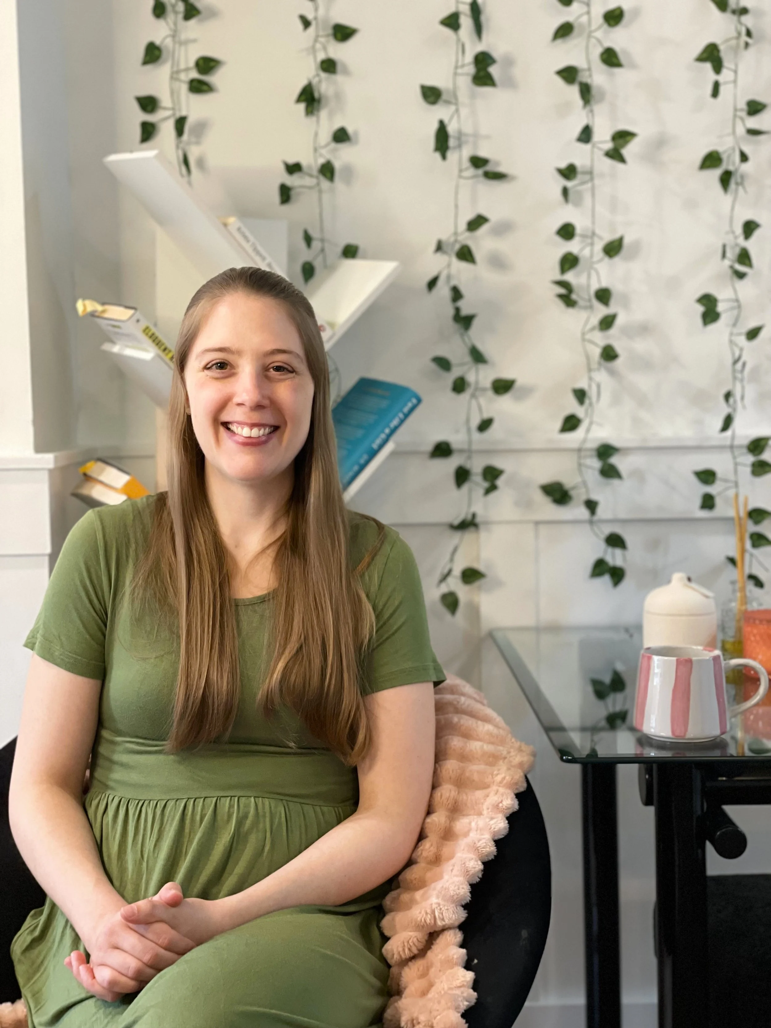 A young woman mental health coach smiling and behind her, there are shelves with books and plants.