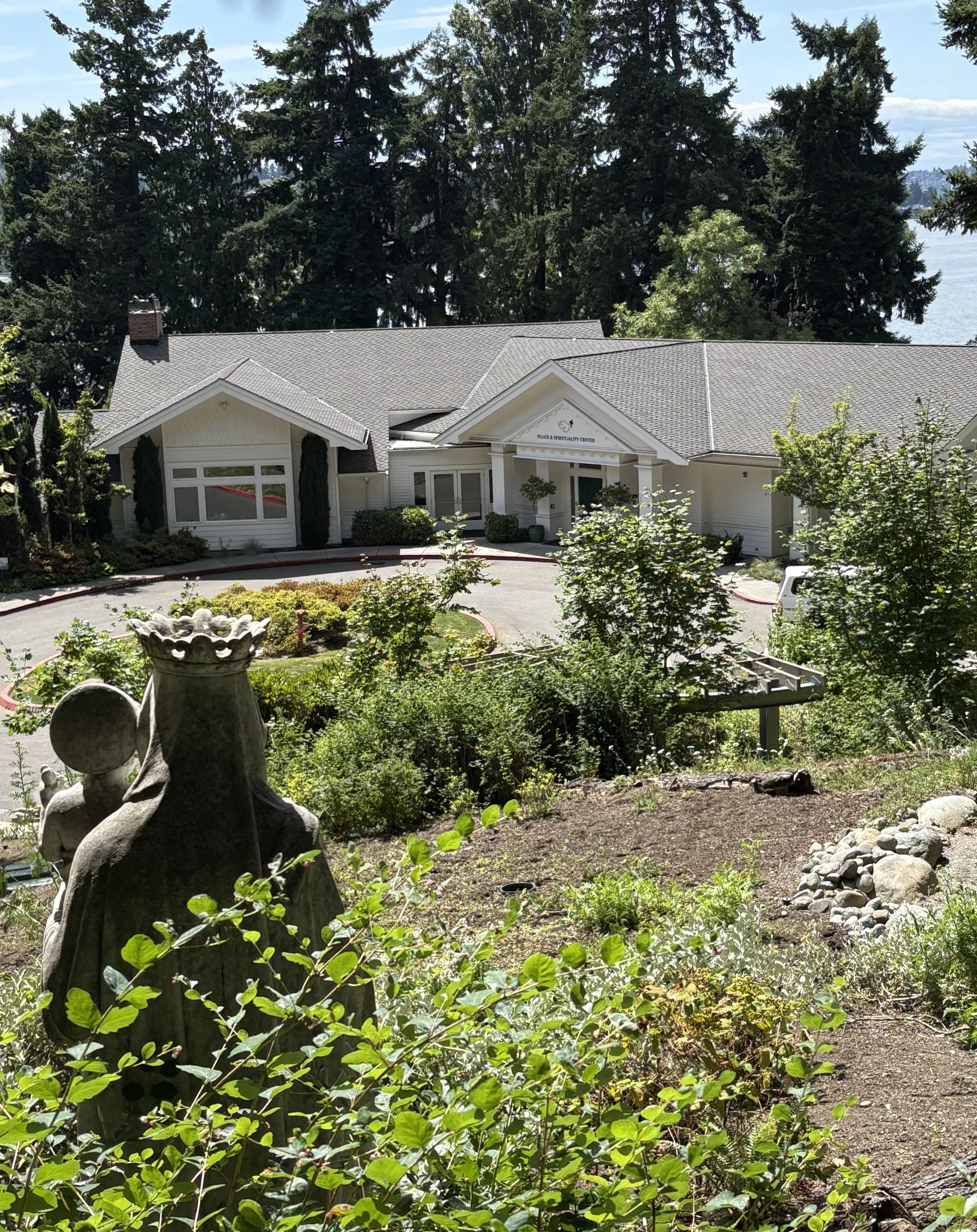 View of a white building labeled 'Place & Spirituality Center' surrounded by trees and greenery, with a garden in the foreground featuring statues and plants; a large tree is visible in the background over the building.