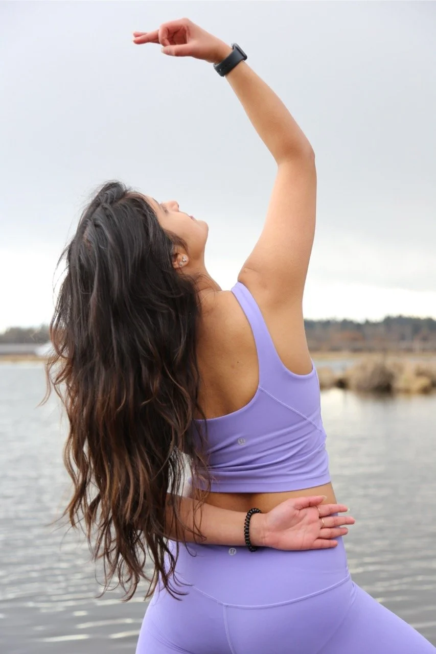 A woman practicing yoga outdoors by a body of water, wearing a purple workout outfit, with her eyes closed and arms raised in a pose.