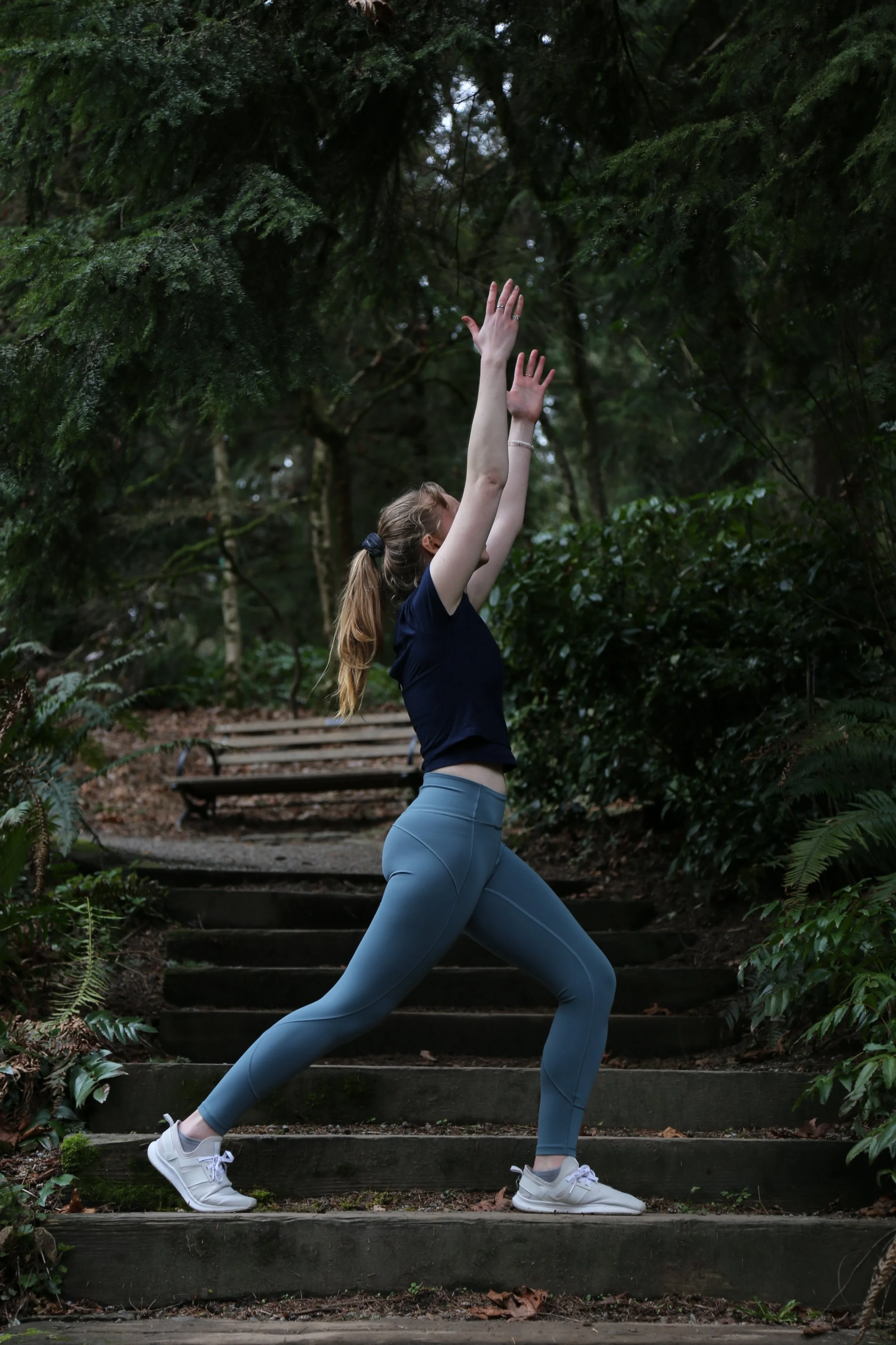 A woman in athletic attire doing a yoga pose on outdoor wooden steps surrounded by lush green foliage.