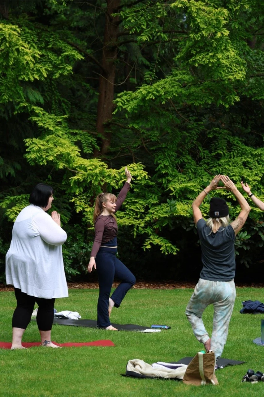 Group  practicing yoga outdoors on a grassy lawn surrounded by green trees.  yoga mats, some standing and doing poses, with one person in the middle raising her arm, and a woman on the left in a white top sitting cross-legged with hands in prayer position.