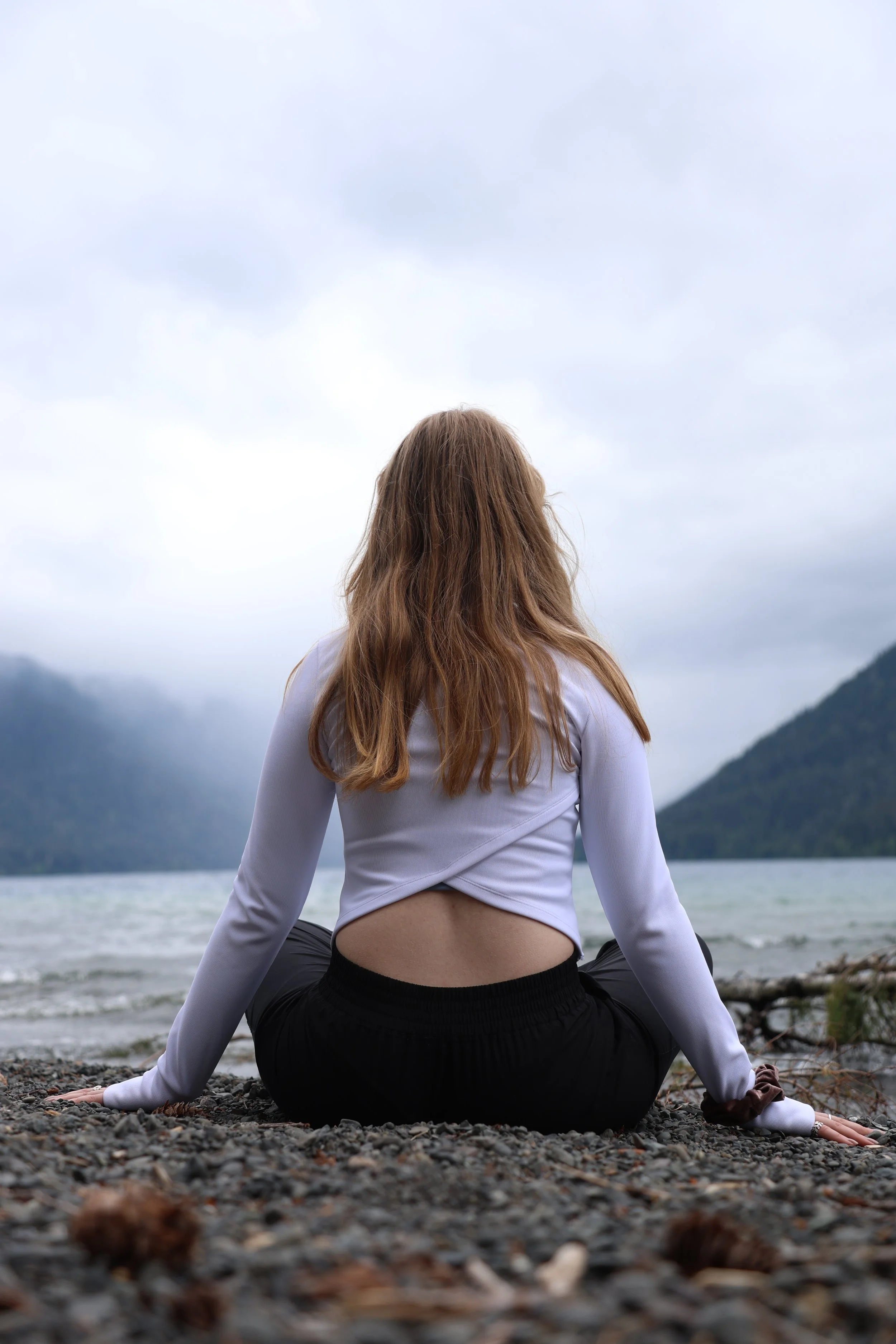A woman sitting in sukhasana and touching the earth on the shore at Lake Crescent, looking out at the water, mountains, and cloudy sky.
