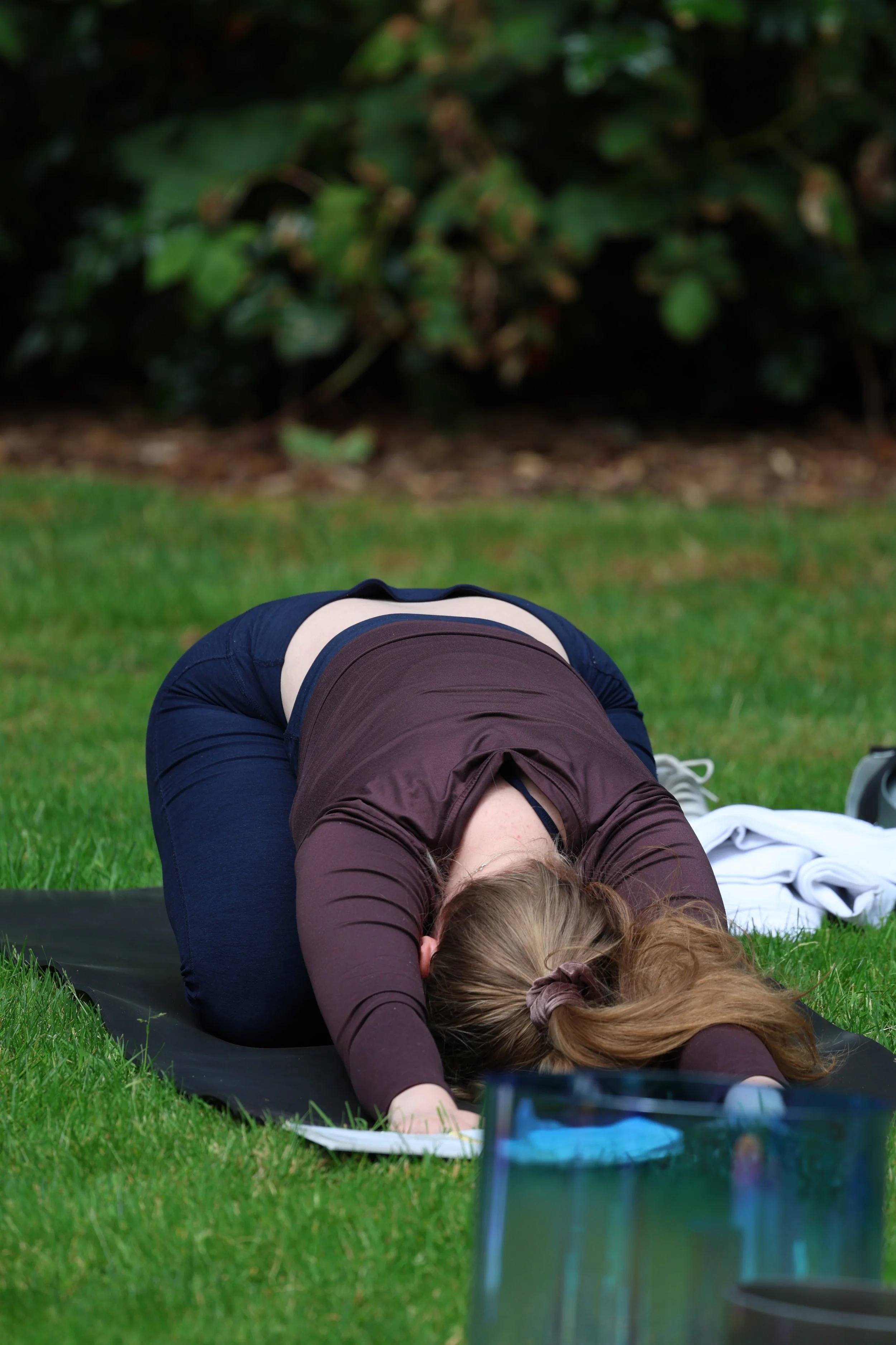 Yoga instructor Zoya practicing yoga outdoors on a grassy lawn, bowing downward in child's pose behind singing bowls that are meant for a sound bath at the end of the practice.