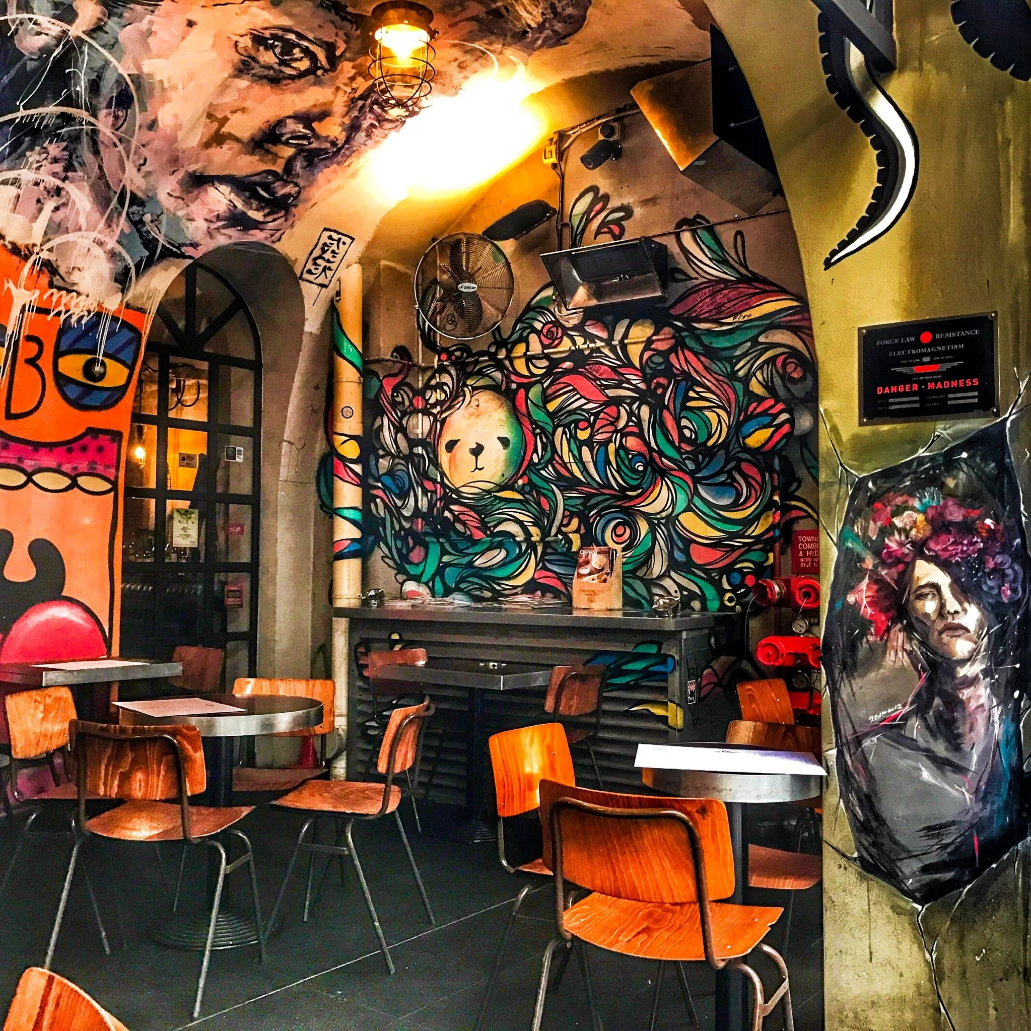 Interior of a cafe or restaurant with colorful wall art, wooden chairs, tables, and various decor elements such as hanging lights, a fan, and paintings.