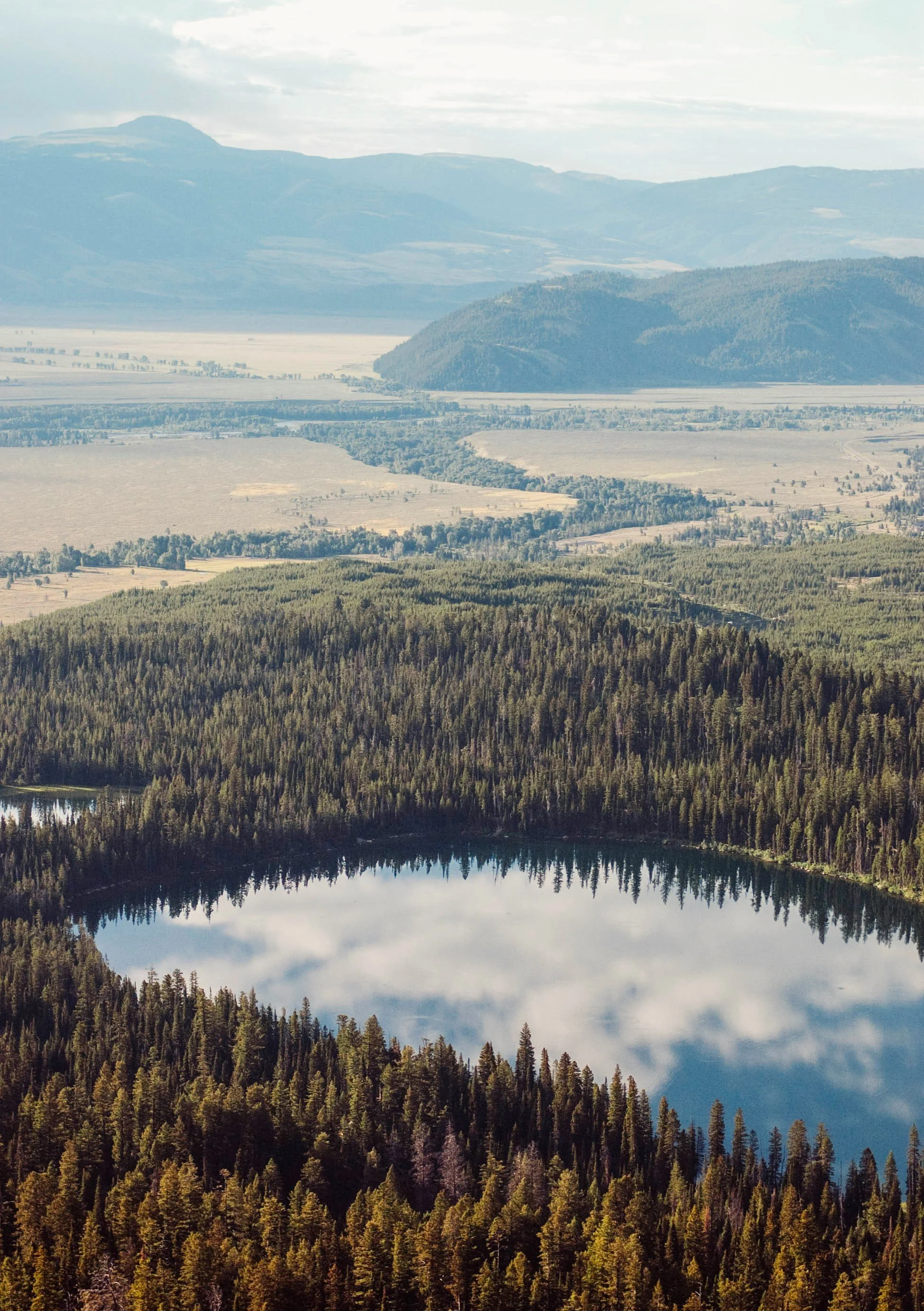 Aerial view of a forested area with a lake reflecting the sky and clouds, surrounded by rolling hills and mountains in the distance.