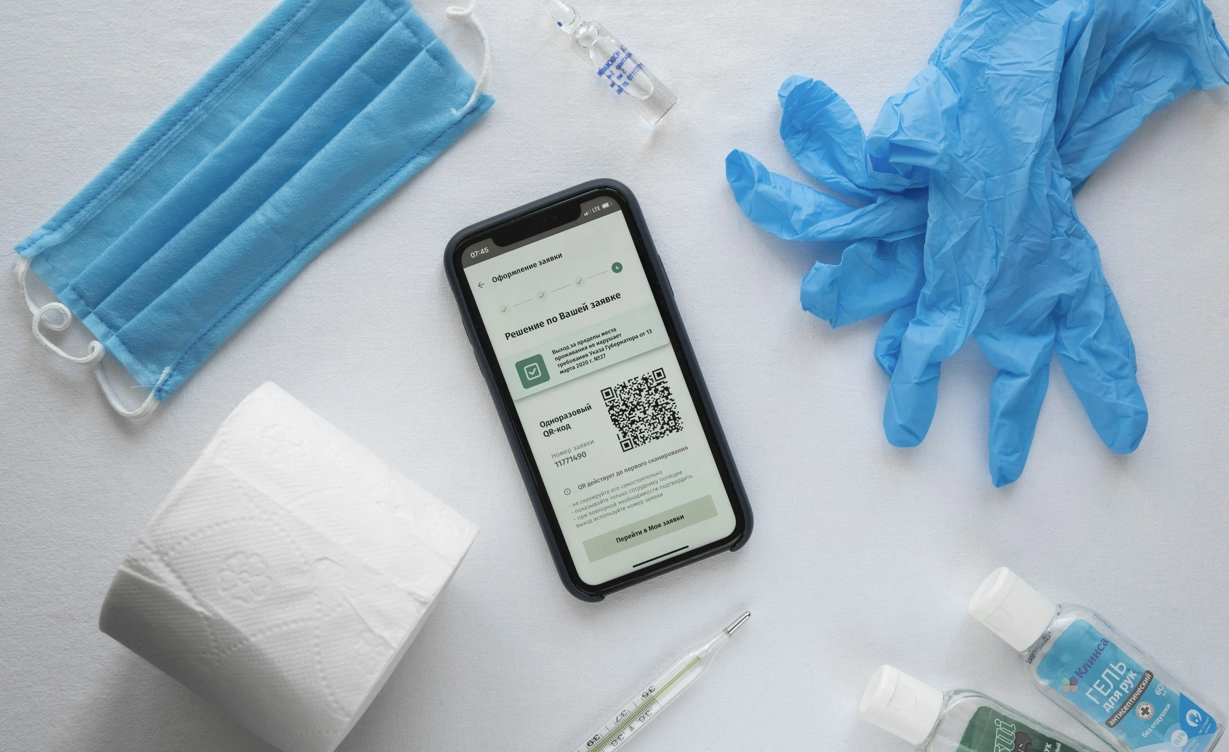A smartphone displaying a QR code with medical and sanitary items around it, including a blue face mask, a roll of toilet paper, blue medical gloves, and bottles of hand sanitizer, on a white surface.