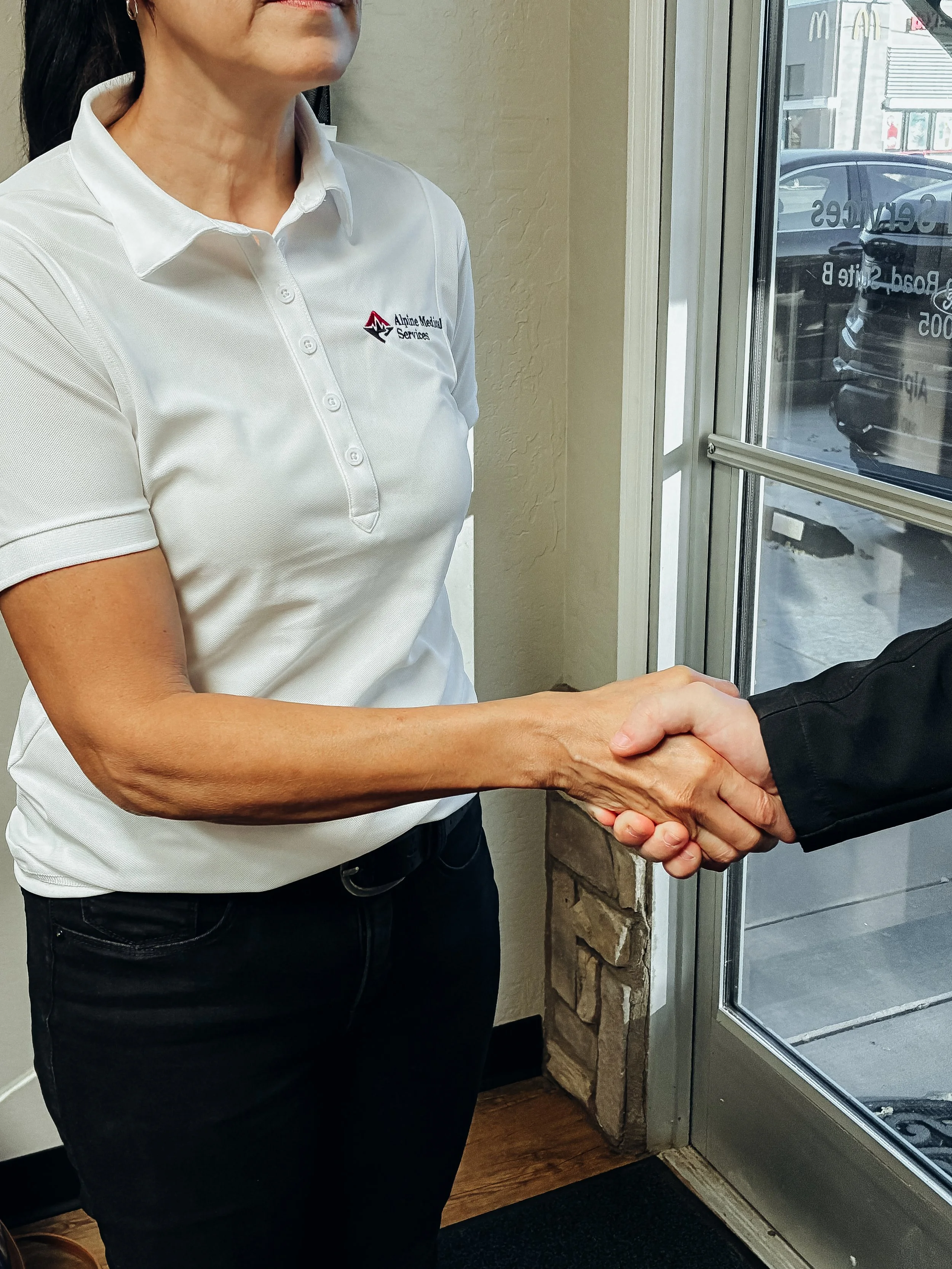 A woman wearing a white polo shirt with a logo shaking hands with a person in a black jacket near a glass door.