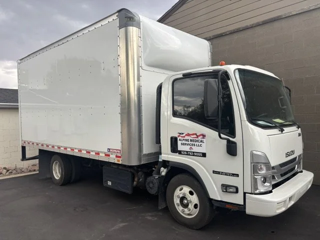 White box truck with a sign for Alpine Medical Services LLC on the door, parked on a paved surface next to a building.