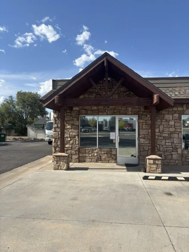 Front of a stone building with a glass door, a porch roof with wooden beams, and a sidewalk in front.