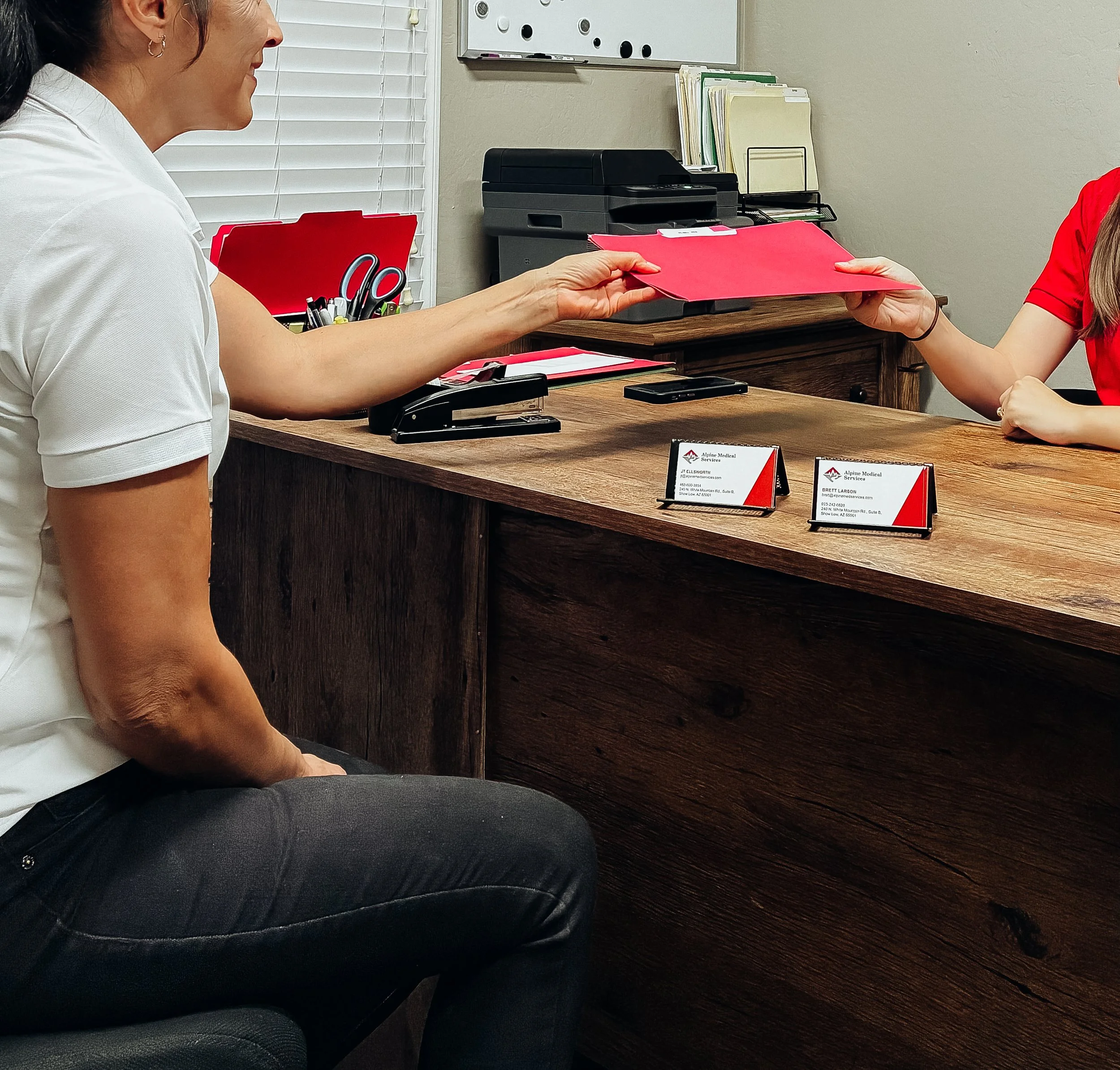 A woman in a white shirt sitting at a wooden desk handing a red folder to a person in a red shirt, with office supplies and nameplates on the desk.