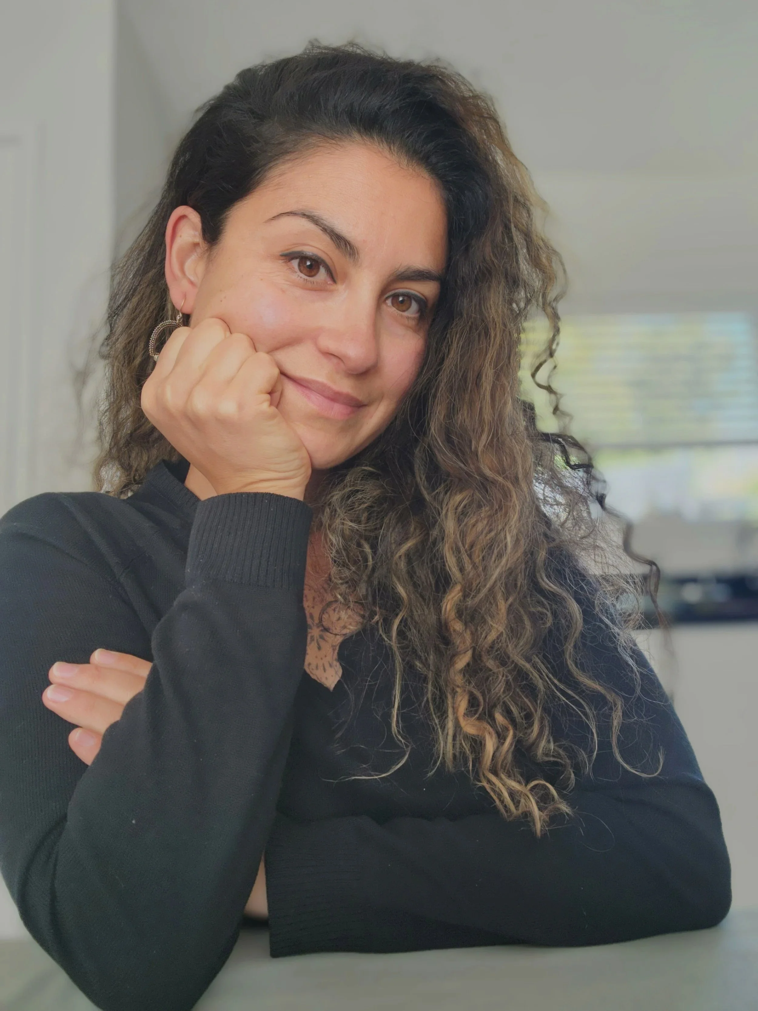 Woman with curly hair resting her chin on her hand, wearing black clothing, sitting at a desk in an indoor setting.