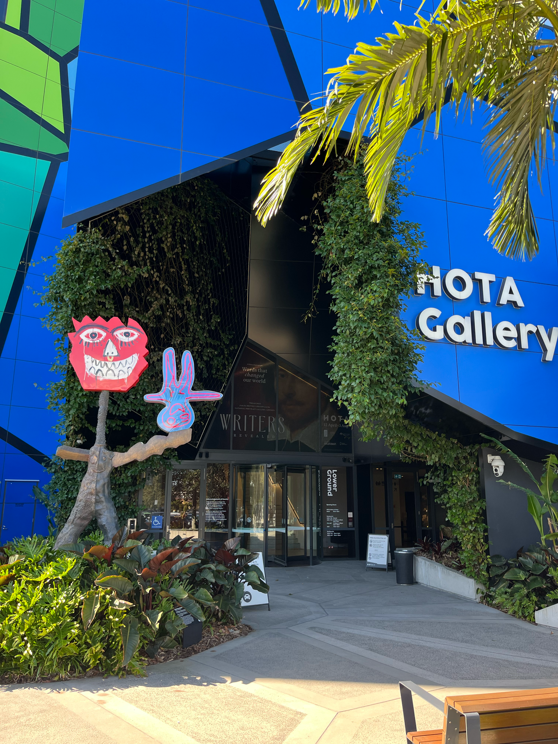 The entrance to the HOTA Gallery building with green plants and colorful art sculptures outside.