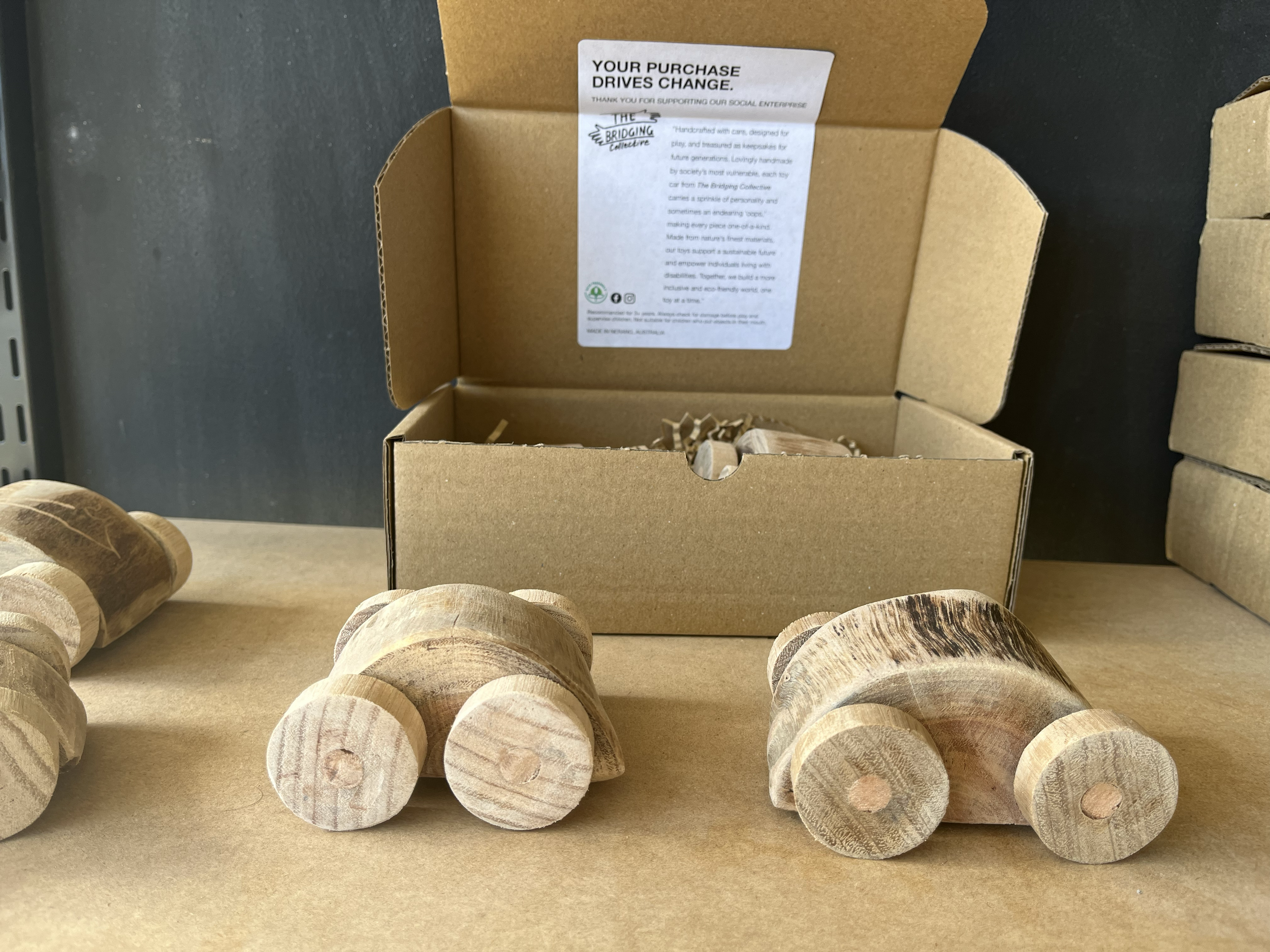 Three small wooden toy cars with rounded wheels placed on a cardboard surface in front of an open cardboard box with a printed label, with stacked boxes to the right.