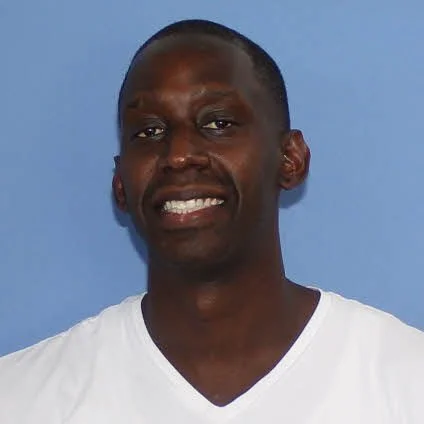 A smiling man with dark skin, short hair, wearing a white shirt against a blue background.