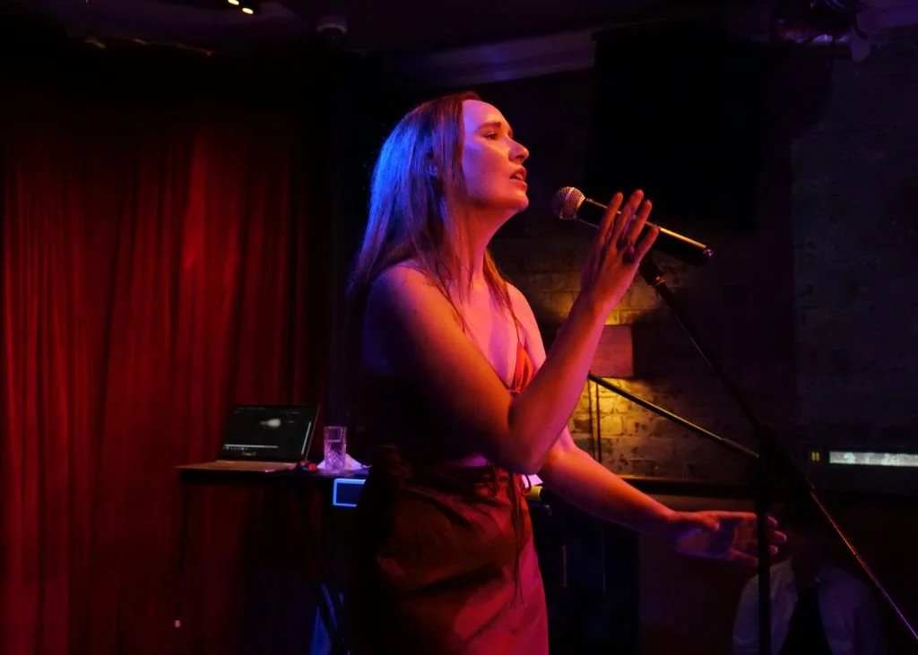 A woman with red hair singing into a microphone on a dimly lit stage with red curtains in the background.