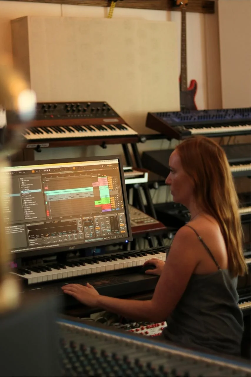 A woman with red hair sitting at a digital audio workstation, working on music production with a computer monitor displaying a digital audio editing software, surrounded by multiple keyboards and musical equipment in a studio.