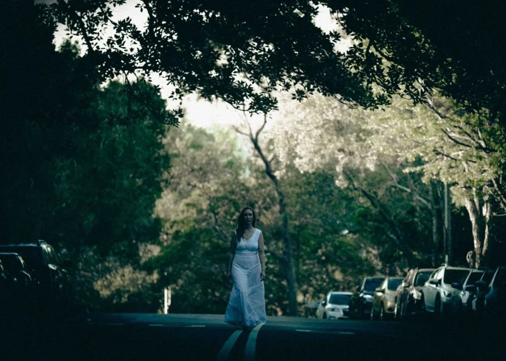 A woman in a white dress walking on a tree-lined street with parked cars on both sides, surrounded by lush green trees, with sunlight filtering through the foliage.