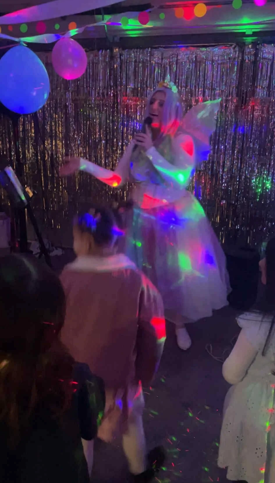 Dazzling woman dressed as a unicorn fairy celebrating at a party with colourful disco lights, balloons, and a shimmering backdrop, singing into a microphone and performing for children.