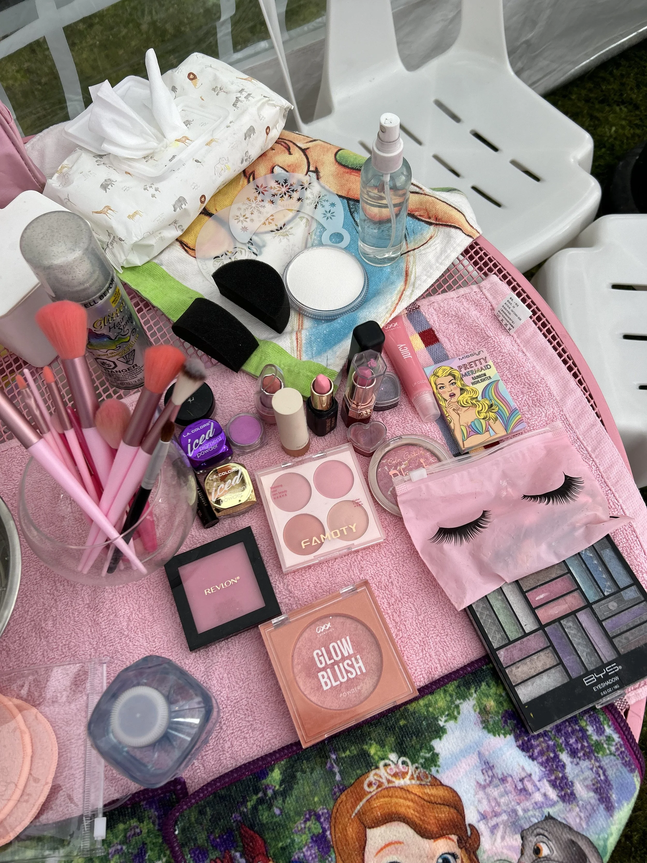 A variety of makeup and beauty products including brushes, eyeshadow, lipstick, false eyelashes, and skincare items, arranged on a pink towel and table with a Disney princess placemat.
