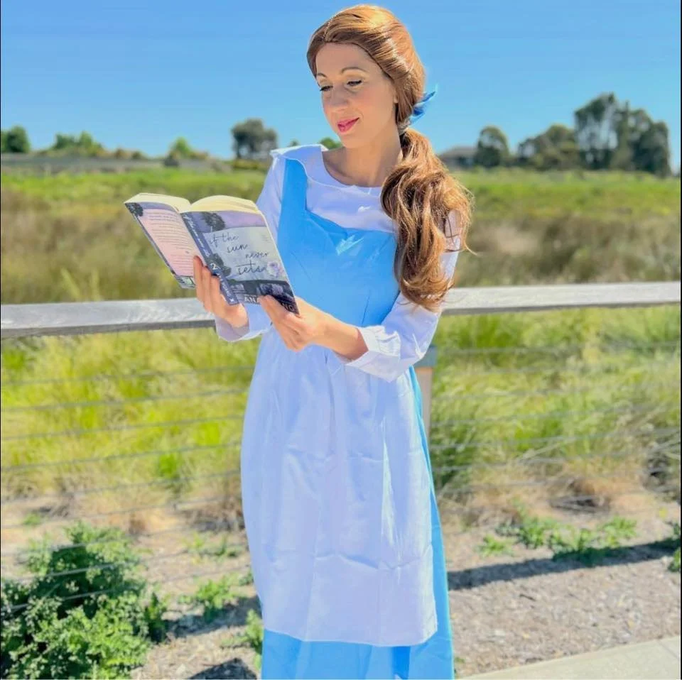 A woman dressed as Peasant Belle from Beauty and The Beast, wearing a blue dress and white apron, is standing outdoors next to a wooden railing, reading a book against a background of green fields and a clear blue sky.