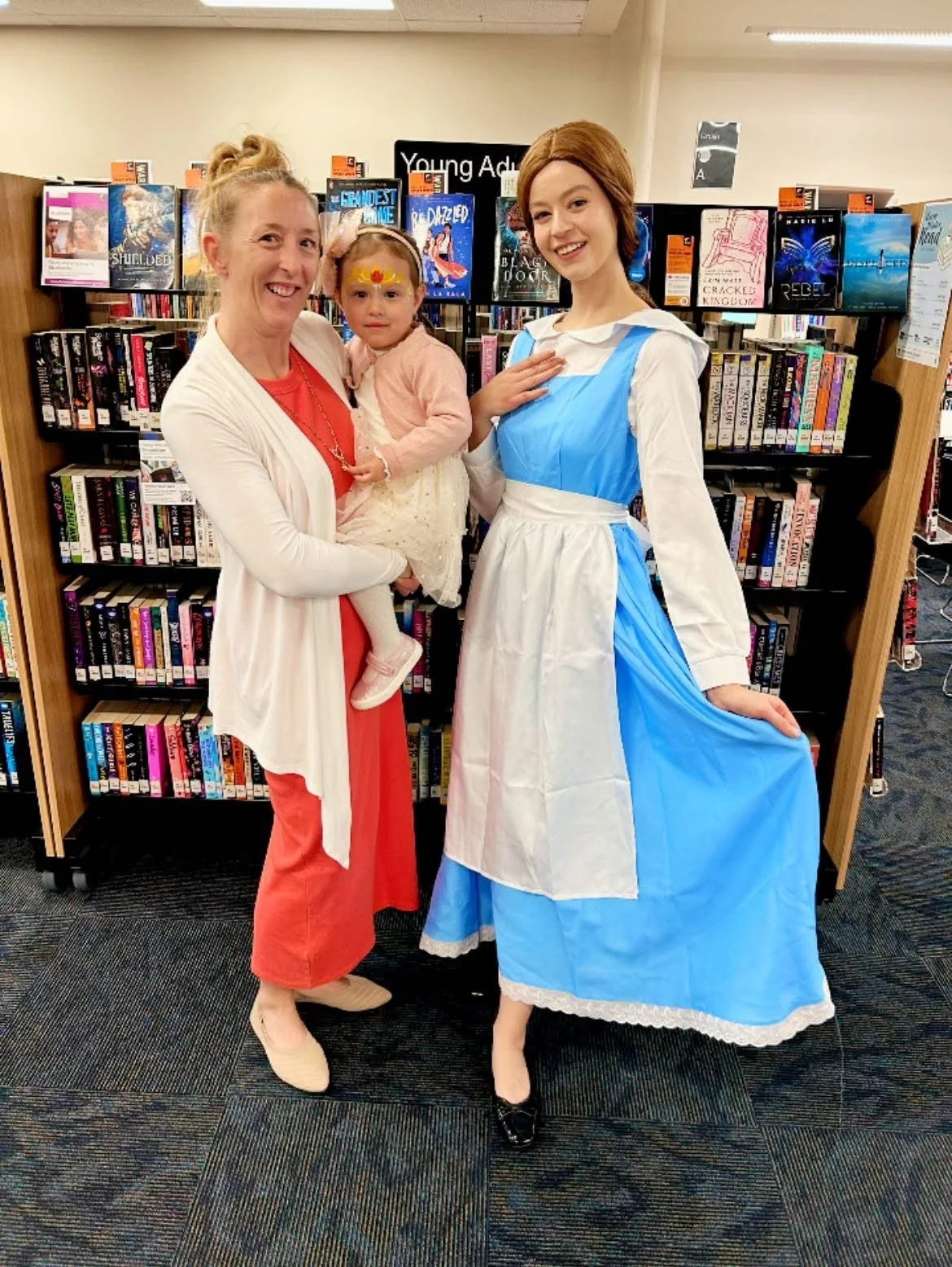 Three females, including a woman, a young girl, and a woman dressed as Belle from Disney's Beauty and the Beast, standing in a library in front of book shelves.