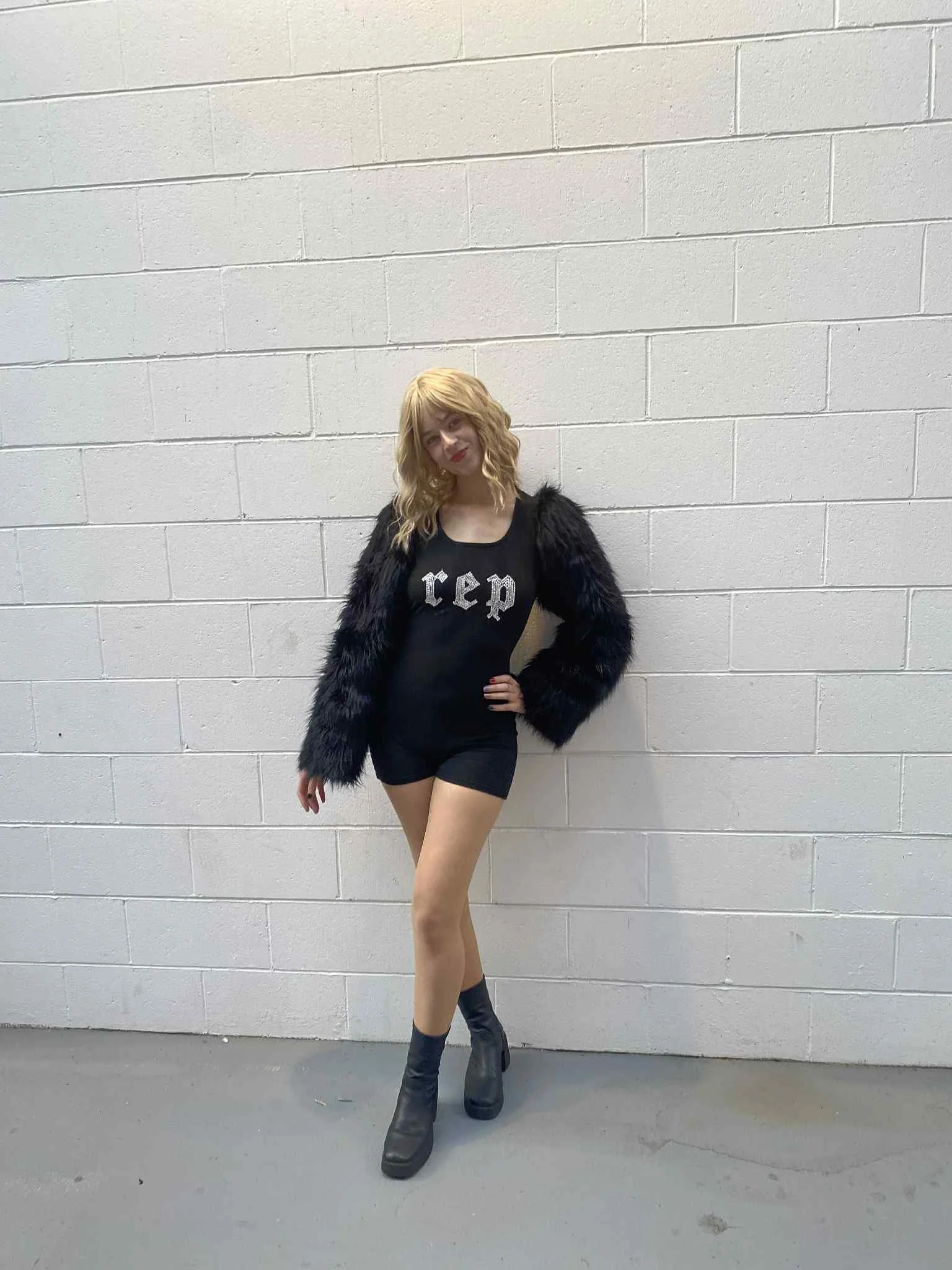 A young woman dressed as Taylor Swift Tay Tay with blonde curly hair posing against a white brick wall wearing a black top and shorts, black fuzzy jacket, and black ankle boots.