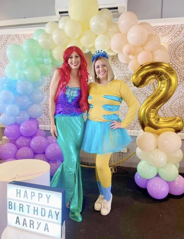 Two women dressed in colorful costumes stand in front of a birthday decoration with rainbow balloons and a large gold balloon shaped as the number 2. One woman wears a mermaid costume with long red hair, purple and blue top, and shiny green fishtail skirt. The other woman wears a yellow and blue clown or fairy outfit with a blue tutu, yellow tights, and a blue tiara. A lightbox sign reads "HAPPY BIRTHDAY AARYA."
