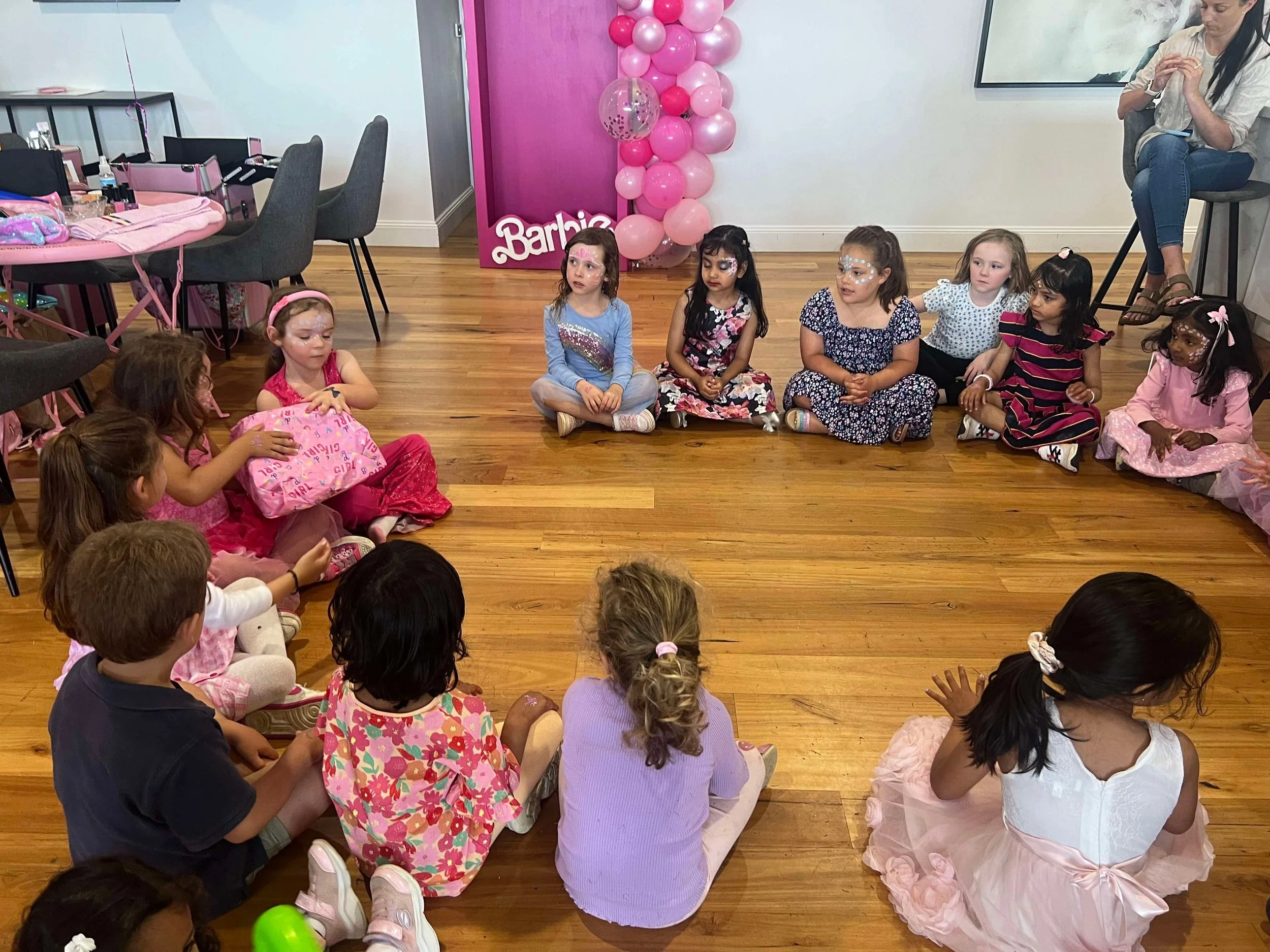 Children sitting in a circle on a wooden floor during a birthday party, with a woman sitting on a stool in the background and pink balloons and a Barbie sign decorating the room.