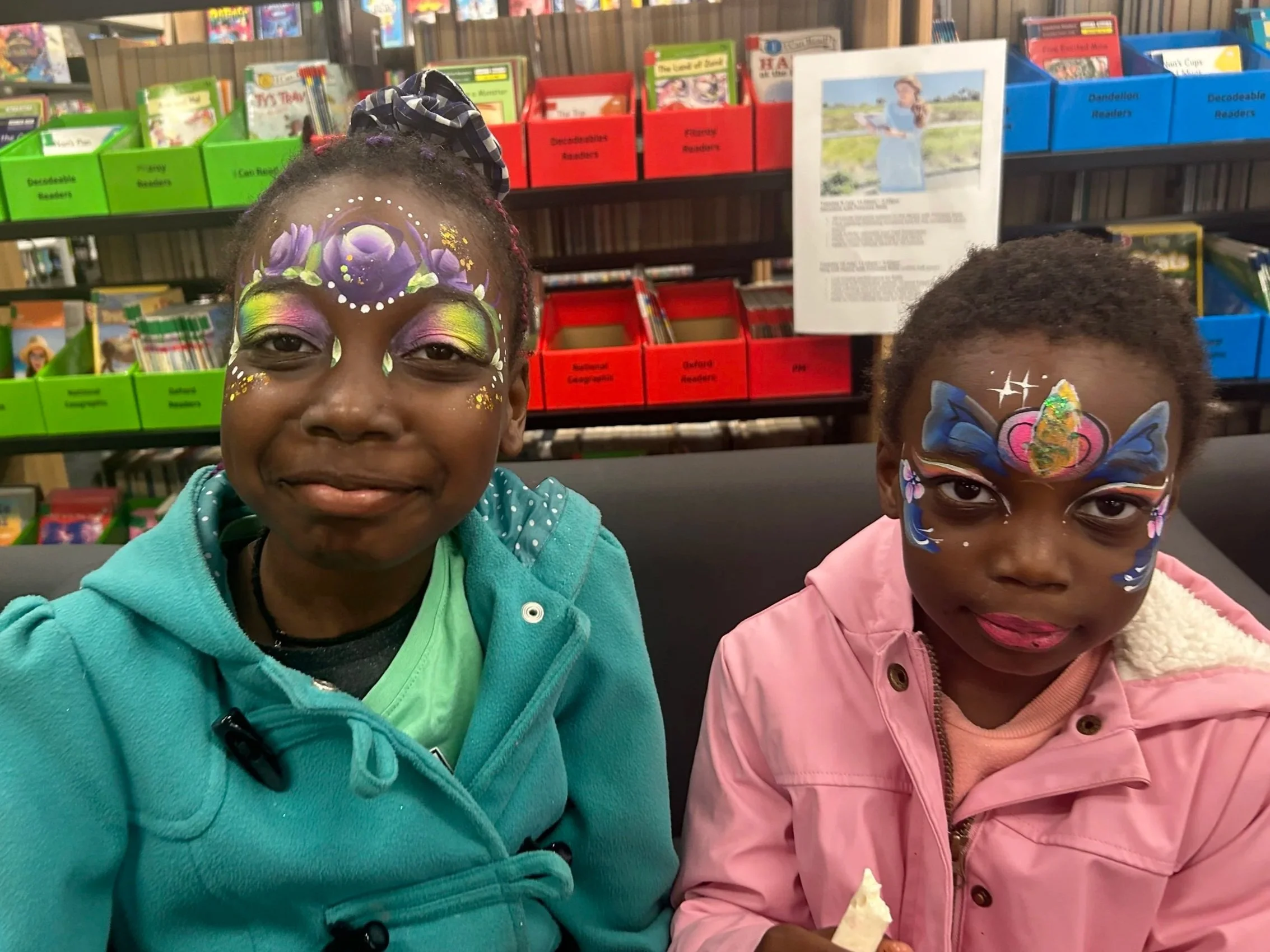 Two children with face paintings, one girl in teal hoodie with floral face art and another girl in pink with butterfly face art, smiling inside a bookstore.