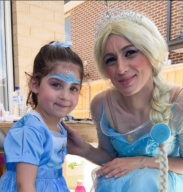 A young girl dressed as a princess and an adult woman dressed as Elsa from Frozen, both smiling, outside on a sunny day.