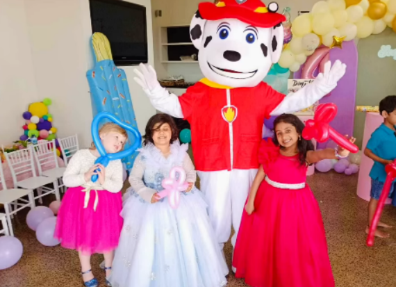 Three young girls and a mascot dressed as Marshall the Dalmatian from Paw Patrol in a red firefighter outfit, posing together at a birthday party with balloons and decorations in the background.