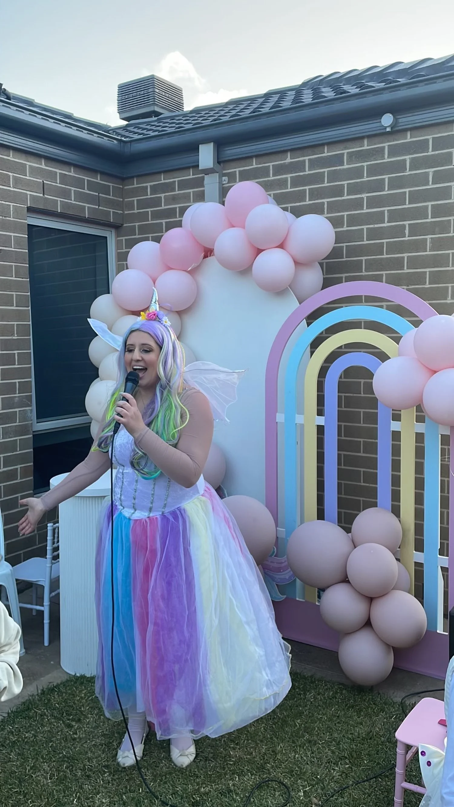 Woman dressed as a unicorn with rainbow-colored hair, fairy wings, and a pastel dress singing into a microphone at a birthday party backdrop decorated with pink balloons and a rainbow arch.