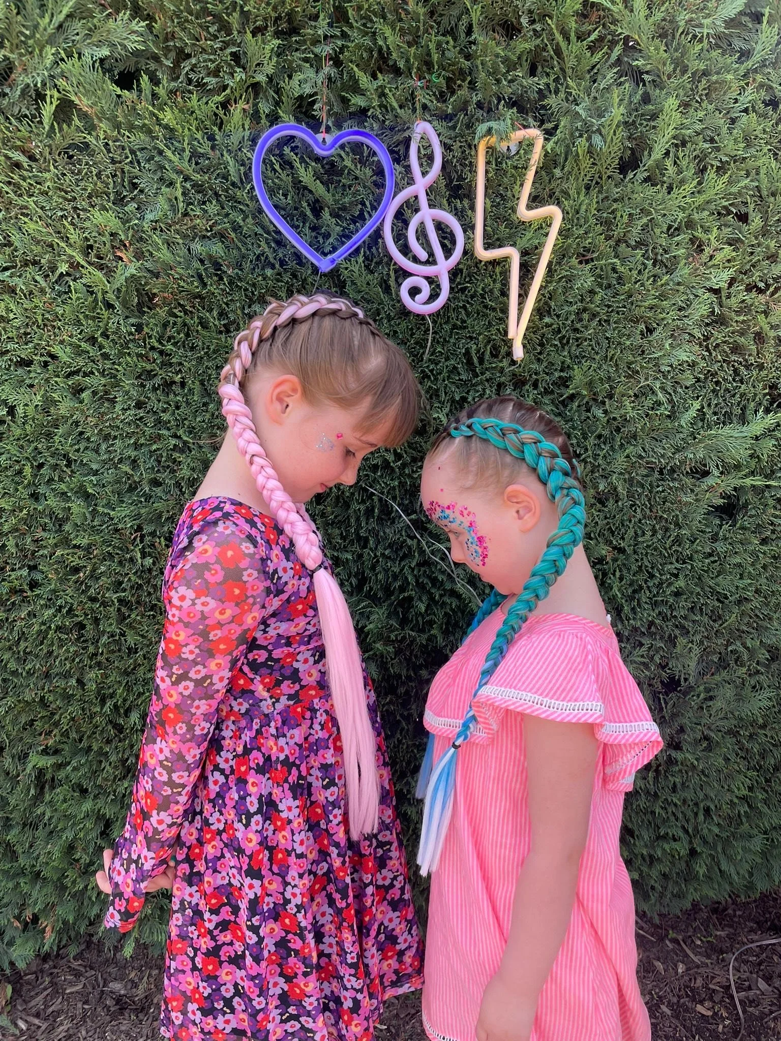 Two young girls with unicorn-braided hair and colourful face paint standing close to each other outdoors, with a green hedge behind them. Neon signs in the background depict a heart, a musical note, a lightning bolt, and a treble clef.