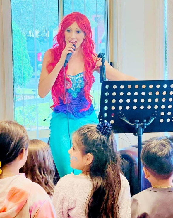 A woman dressed as a Ariel the mermaid from Disney's The Little Mermaid with bright red hair, singing into a microphone for children in a room with large windows.