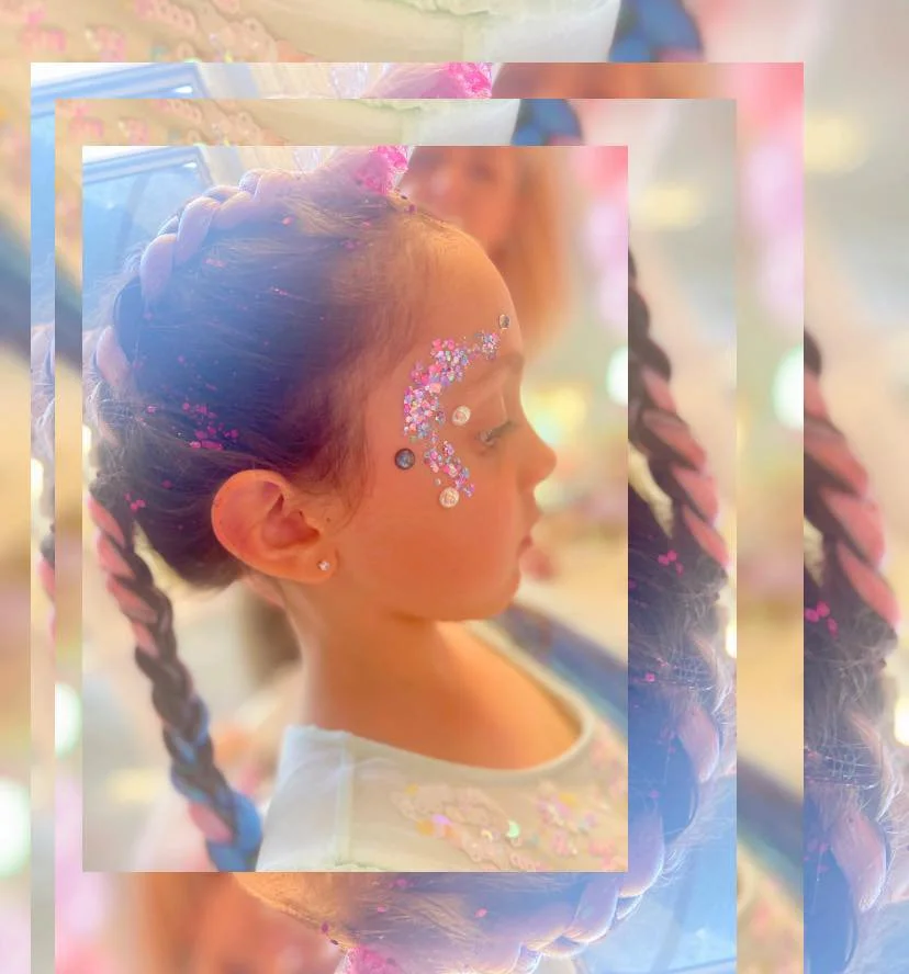 A young girl with her hair styled in a braid, decorated with pink glitter and rhinestones on her face, in a festive setting.