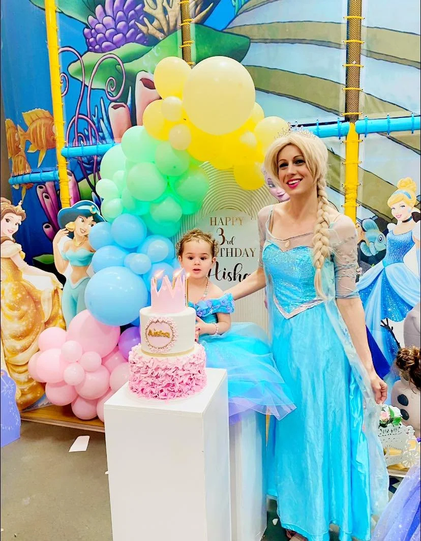 A young girl dressed as a princess, sitting at a birthday celebration with a woman dressed as Elsa from Frozen, with colorful balloons and a themed backdrop.