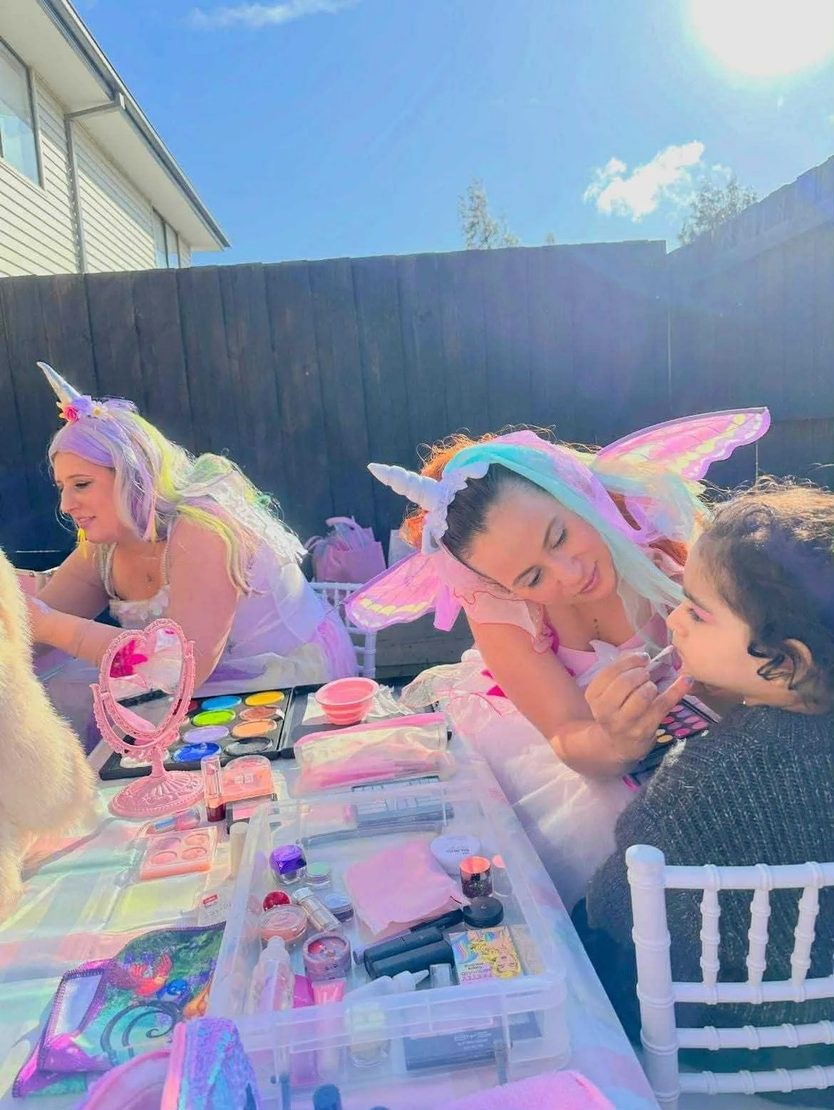 Children dressed in unicorn and fairy costumes having face paint at a birthday party outdoors on a sunny day.