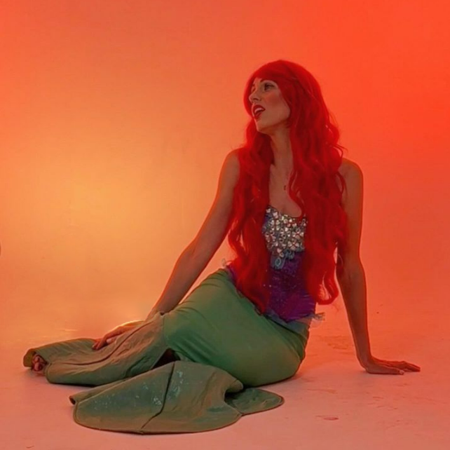A woman with long red hair sitting on the floor in front of a gradient orange background. She is wearing a sequined top and flowing green pants, looking to her right with an expressive face.