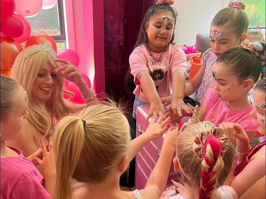 A group of young girls with face paint and glitter, gathered around a woman wearing a tiara, playing a game at a birthday party. The background has pink and red balloons and decorations.