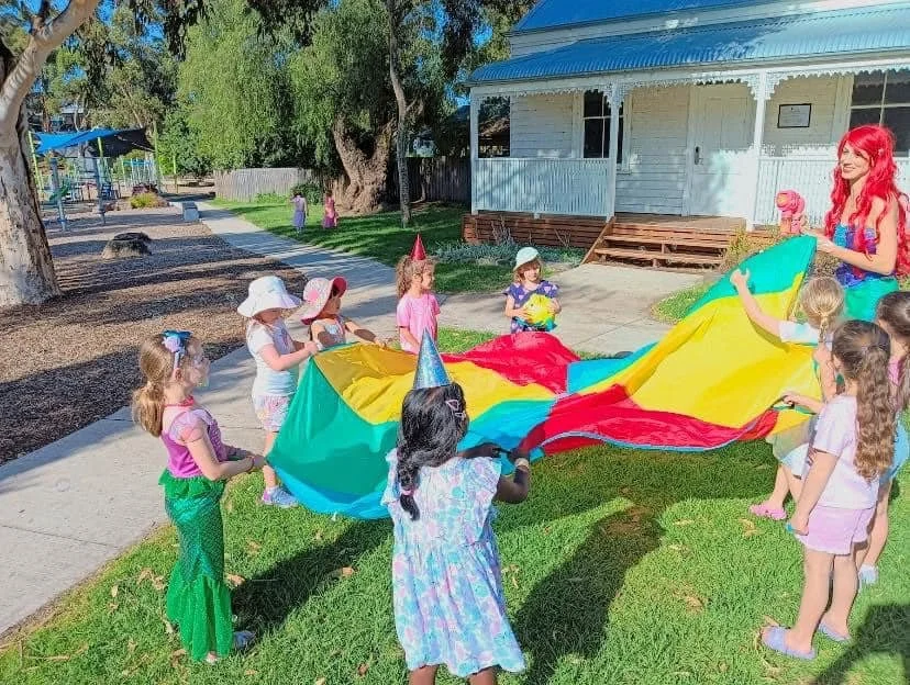 Children celebrating a birthday party outdoors with a woman dressed as Ariel from The Little Mermaid, playing with a colorful parachute.