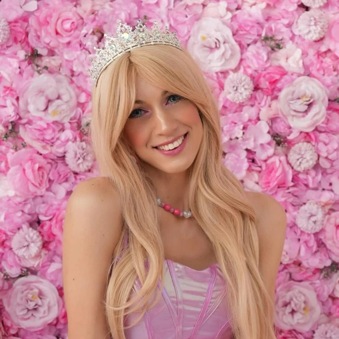 A young woman dressed as Barbie with long, wavy blonde hair wearing a silver tiara and a pink dress, standing in front of a pink flower wall.