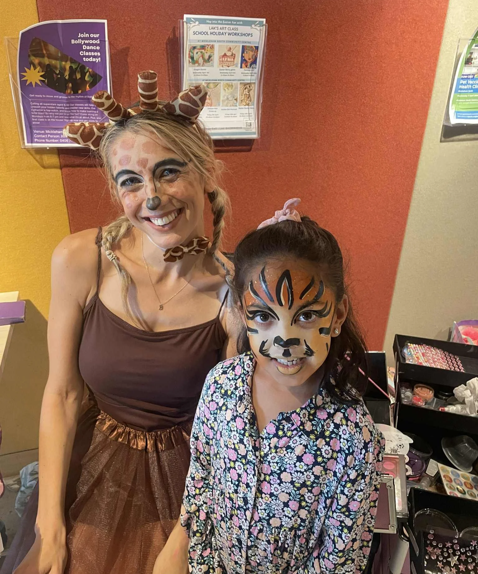 Two smiling women with animal face paint, one dressed as a giraffe and the other as a tiger, at a festive event with face painting supplies in the background.