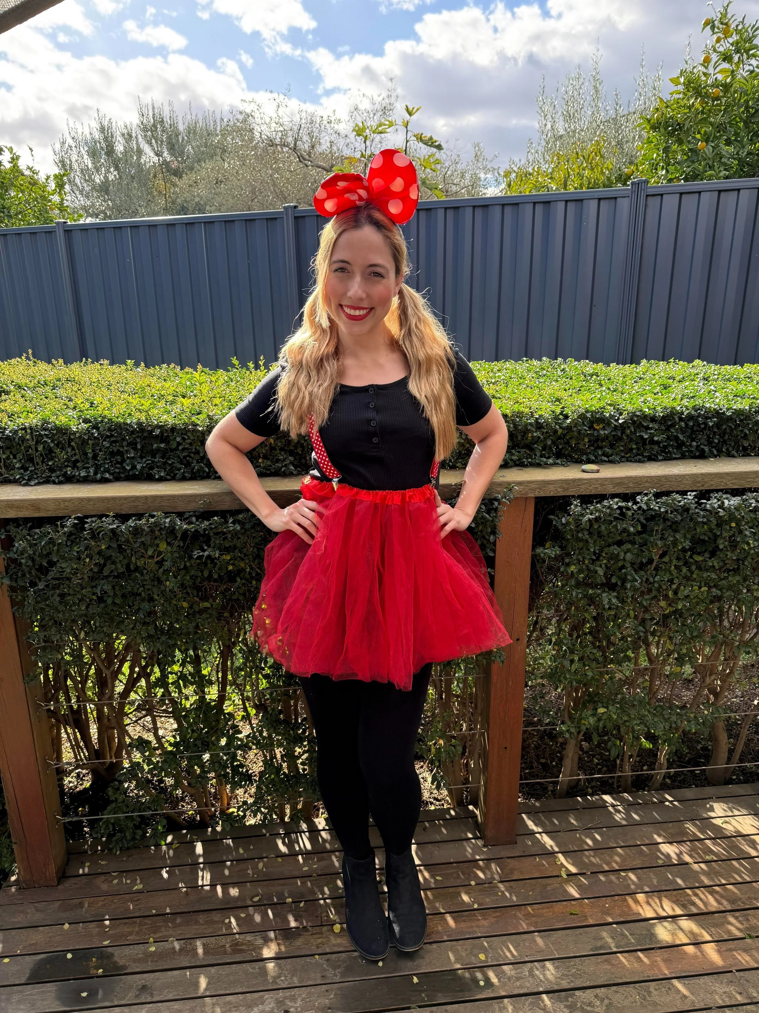 Young woman in a Minnie Mouse-inspired costume with a red polka-dot bow, red tutu, black top, black leggings, and black boots, standing outdoors on a wooden deck with a garden and blue fence in the background.
